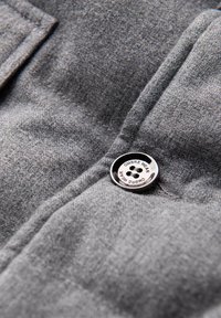 Tejido gris con una textura suave, que presenta costuras visibles y un botón negro redondo etiquetado como "OMBRE WEAR" con cuatro agujeros.