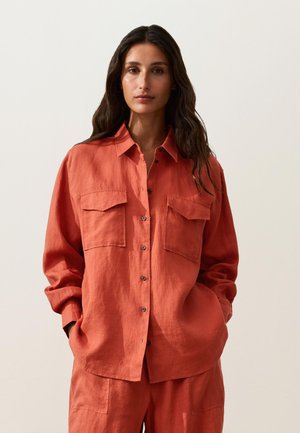 Woman with long dark hair wearing a loose-fitting burnt orange button-up shirt and matching pants, standing with hands in pockets.
