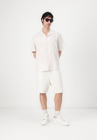 Κοντομάνικο πουκάμισο με κουμπιά σε ανοιχτό μπεζ χρώμα με κάθετες ρίγες, συνδυασμένο με λευκές υφασμάτινες βερμούδες και λευκά αθλητικά παπούτσια. Μαύρα γυαλιά ηλίου.