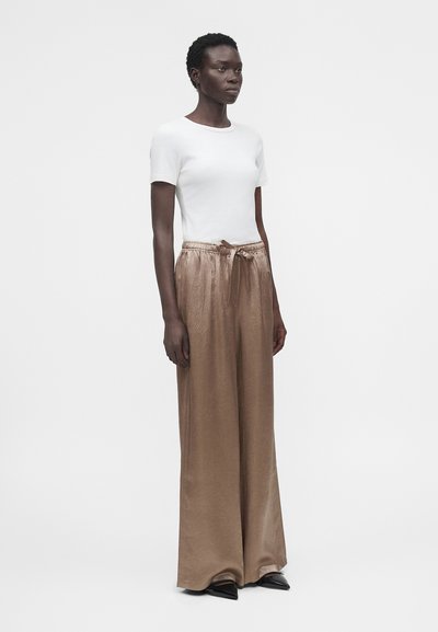 Γυναίκα που φοράει εφαρμοστό λευκό κοντομάνικο μπλουζάκι και φαρδιά, ψηλόμεσα μπεζ παντελόνια με μαύρα μυτερά παπούτσια.