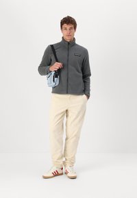 Giacca in fleece grigia con zip, pantaloni in velluto a coste beige e sneakers color crema con strisce rosse. Tiene una borsa a tracolla azzurro chiaro.