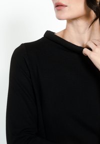 Črn pulover z visokim, zloženim ovratnikom, dolgimi rokavi in gladkim, teksturiranim materialom. Posnetek v povezavi z ovratnikom in področjem ramen.