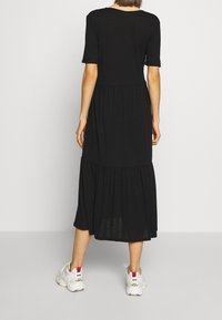 Schwarzes knielanges Kleid mit kurzen Ärmeln, gestuftem Rockdesign und entspannter Passform. Getragen mit weißen Sneakers mit roten Akzenten.