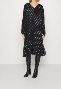 Robe noire avec un décolleté en V, des manches longues et des motifs ovales argentés. Tissu fluide et coupe évasée, associée à des bottes noires arrivant aux genoux.