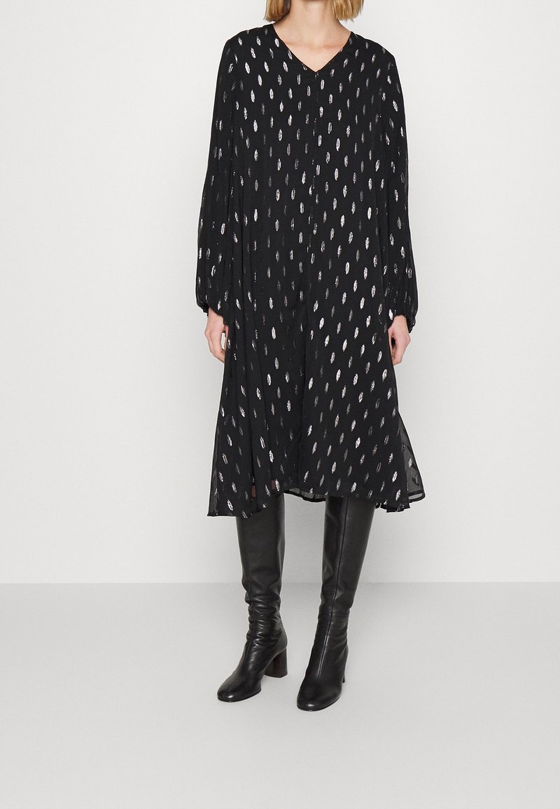 Robe noire avec un décolleté en V, des manches longues et des motifs ovales argentés. Tissu fluide et coupe évasée, associée à des bottes noires arrivant aux genoux.