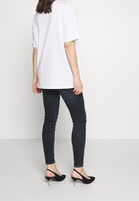 Una mujer de pie, de espaldas, vistiendo una camiseta blanca holgada, jeans oscuros ajustados y tacones negros tipo slingback, frente a un fondo blanco liso.