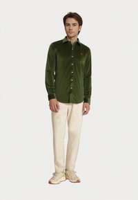 Giovane uomo che indossa una camicia a maniche lunghe verde scuro, pantaloni beige e sneakers chiare, in piedi contro uno sfondo bianco uniforme.