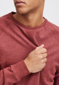 Sweat-shirt bordeaux avec un col rond et des manches raglan, présentant une texture douce et des cuffs côtelés. Le tissu a un aspect légèrement chiné.