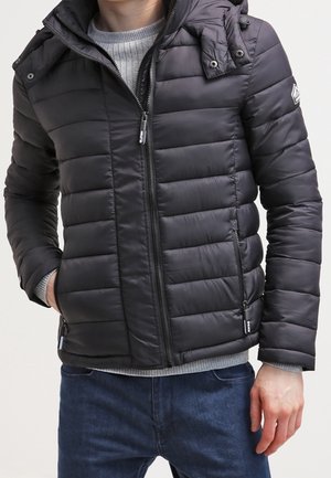 Mand iført en sort quiltet jakke med lynlås foran og lynlåslommer over en grå sweater og blå jeans.