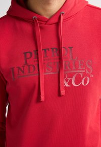 Hanorac roșu cu textul logo „PETROL INDUSTRIES & Co” pe față și șnururi de aceeași culoare, purtat de o persoană decupată din gât în jos.