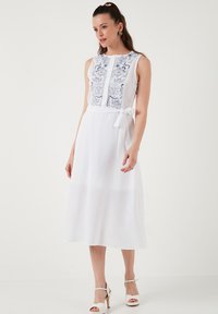 Weißes ärmelloses Kleid mit bestickten blauen Details, Knopfleiste und gebundenem Taillenband. Es hat eine mittellange, leichte Leinenqualität.