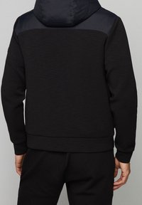 Hombre vestido con una sudadera negra que tiene un panel más oscuro en la parte superior de la espalda y la capucha, combinado con pantalones negros, visto desde atrás.