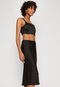 Schwarzes Satin-Crop-Top mit asymmetrischem Design und dünnen Trägern, kombiniert mit einem passenden schwarzen Satin-Midi-Rock, der eine glatte Textur aufweist.