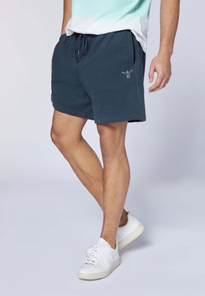 Mann trägt marineblaue Shorts mit Kordelzug und kleinem gesticktem Logo, weiße Turnschuhe und ein Teil eines hellblauen und weißen Hemds sichtbar.