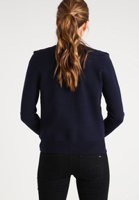 Granatowy sweter z dzianiny o gładkiej fakturze, dopasowanym kroju i ozdobach na ramionach. Widok z tyłu pokazuje ściągany dół oraz dopasowane rękawy.