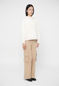 Ženska stoji oblečena v belo kapuco z detajlom cvetlice in bež široke cargo hlače ter črne čevlje na belem ozadju.