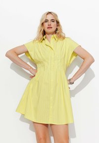 Femme aux cheveux blonds portant une robe chemise jaune à manches courtes avec de fines rayures verticales, les mains sur les hanches, se tenant devant un fond blanc.