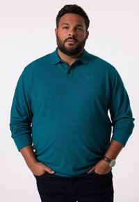 Teal långärmad polo med knapplock, med en subtil logotyp på bröstet och en slät textur. Bärs tillsammans med mörka jeans.