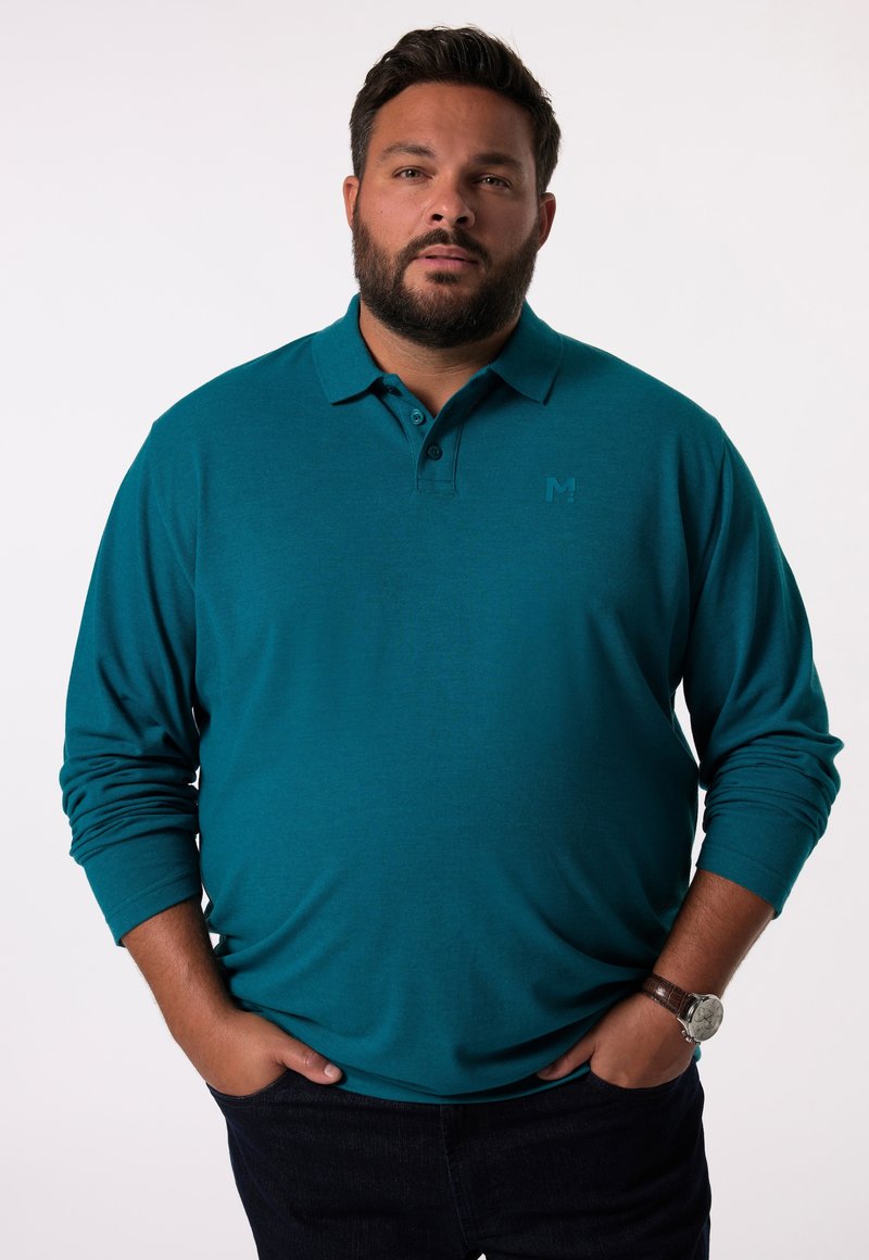 Teal långärmad polo med knapplock, med en subtil logotyp på bröstet och en slät textur. Bärs tillsammans med mörka jeans.