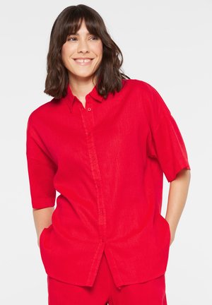 Frau mit dunklen Haaren, lächelnd, trägt ein leuchtend rotes Kurzarmhemd mit Knöpfen und passende rote Hose, die Hände in den Taschen.
