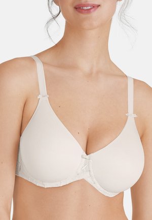 Kvinde iført en beige undertrusse-bh med tynde justerbare stropper og små dekorative sløjfer nær stroppene og midten.