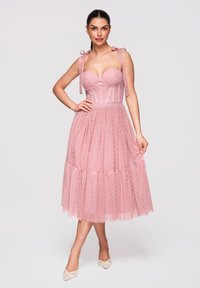 Femme en robe midi en tulle rose sans manches à pois avec bretelles en nœud, debout avec une main sur la hanche et des talons blancs à bout pointu.
