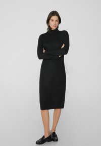 Schwarzes gestricktes Kleid mit hohem Kragen, langen Ärmeln und gerader Silhouette, das bis zur Mitte der Waden reicht. Kombiniert mit schwarzen Slippern.