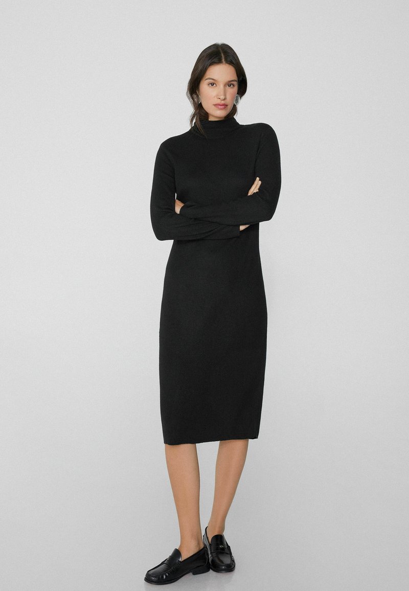 Schwarzes gestricktes Kleid mit hohem Kragen, langen Ärmeln und gerader Silhouette, das bis zur Mitte der Waden reicht. Kombiniert mit schwarzen Slippern.