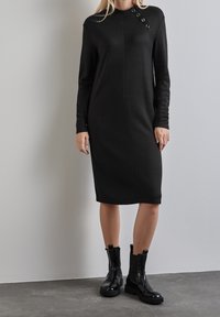 Robe noire à manches longues avec un col haut et un accent de boutons, fabriquée à partir d'un tissu lisse. Associée à des bottines noires à semelles épaisses.