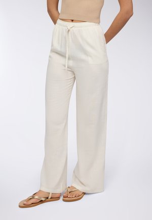 Pantaloni crema leggeri e larghi con una fascia elastica in vita e cordoncino. Texture liscia, gambe dritte, abbinati a infradito beige.