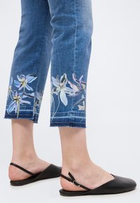 Κατώτερα πόδια με μπλε cropped τζιν με φλοράλ σχέδια κοντά στα τελειώματα και μαύρα φλατ παπούτσια slingback σε λευκό φόντο.