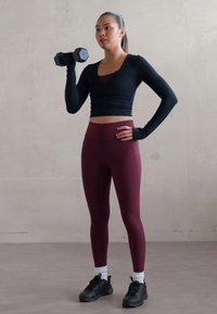 Zwarte lange mouwen cropped top, bordeaux leggings en zwarte sneakers. Houdt een dumbbell in de rechterhand, staat met de linkerhand op de heup.