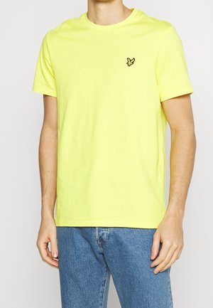 Mężczyzna ubrany w jaskrawożółty t-shirt z krótkim rękawem, z małym czarnym logo ptaka na lewej piersi, oraz w niebieskie jeansy.