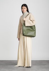 Green textured bag with a floral pattern, carried by a model wearing a beige oversized sweater and cream wide-leg trousers.
