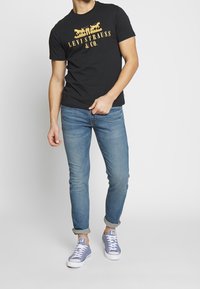 Schwarzes Baumwoll-T-Shirt mit gelbem Text und Grafik, kombiniert mit hellblauen Jeans und grauen Leinenschuhen, vor einfachem Hintergrund stehend.