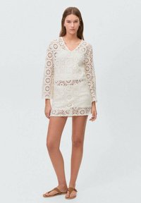 Weißes Häkelkleid mit langen Ärmeln, das hexagonale und florale Muster aufweist. Leichter Stoff, knielang, kombiniert mit braunen Sandalen.