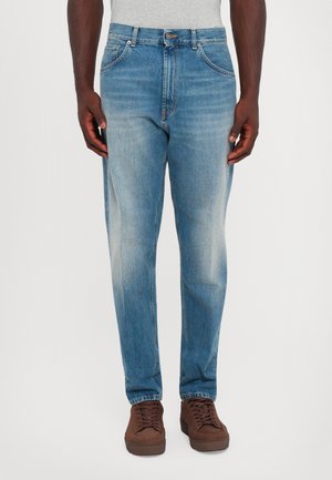 Uomo che indossa jeans sfumati blu chiaro e scarpe marroni con lacci, in piedi di fronte a uno sfondo bianco uniforme.