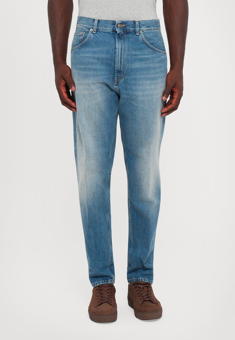 Uomo che indossa jeans sfumati blu chiaro e scarpe marroni con lacci, in piedi di fronte a uno sfondo bianco uniforme.
