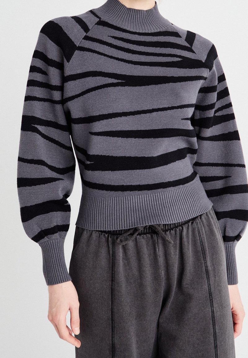 Pull gris tricoté avec des rayures zèbre noires, doté d'un col montant et de poignets côtelés. Associé à un pantalon foncé à coupe décontractée.