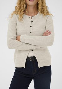 Šviesiai pilkas kardiganas iš tekstūruotos megztos medžiagos, su apvalia iškirpte, juodais mygtukais ir ribinėmis rankogaliais, derinamas su tamsiomis džinsais.