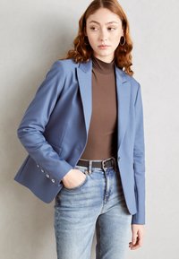 Modrý blazer z hladké látky s klopami, jedním knoflíkem a detaily na manžetách se stříbrným knoflíkem, v kombinaci s hnědým topem a vybledlými džíny.