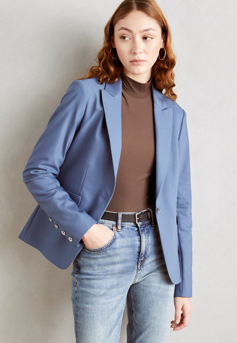 Modrý blazer z hladké látky s klopami, jedním knoflíkem a detaily na manžetách se stříbrným knoflíkem, v kombinaci s hnědým topem a vybledlými džíny.