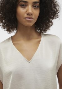 Haut beige clair à col en V avec des manches courtes. Tissu lisse, légèrement texturé avec un éclat subtil et un détail de couture ton sur ton.