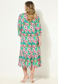 Blumiges Midikleid mit grünem Hintergrund, verziert mit rosa Hibiskusblüten und Schmetterlingen. Mit einem geschichteten Rock und Dreiviertelärmeln. Silberne Absätze.