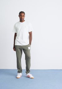 Tânăr stând în interior îmbrăcat cu un tricou alb, pantaloni de trening verde măslin cu o patroană și teniși albi cu dungi roșii.