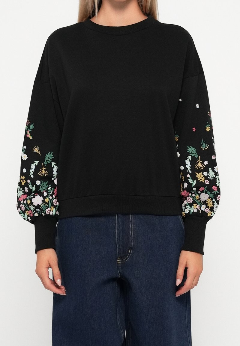 Top noir à manches longues avec des motifs floraux colorés sur les manches bouffantes, porté avec un jean bleu foncé, visible du cou jusqu'aux hanches.
