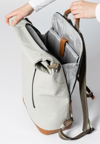 Zaino grigio chiaro con dettagli in pelle marrone, dotato di chiusura a risvolto e fodera grigia a motivo. Spallacci regolabili e tasca laterale con zip.