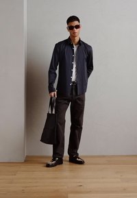Camicia nera a bottoni, t-shirt bianca, pantaloni scuri e scarpe nere lucide. Gli accessori includono occhiali da sole marroni e una borsa a tracolla nera.
