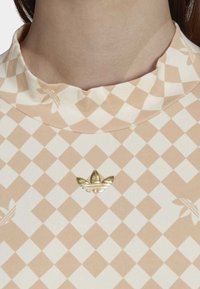 Hřejivá a krémová kostkovaná blůza s vysokým límcem, která má na středu zlaté kovové logo a jemné květinové vzory.
