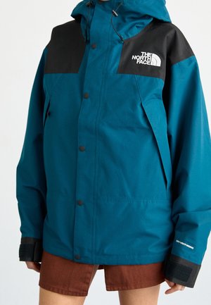 Veste imperméable The North Face bleu sarcelle et noire avec capuche, boutons à l’avant et poignets velcro portée avec un short marron.